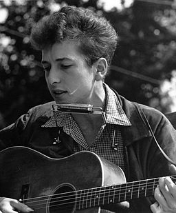 A black and white photo of young Bob Dylan with an acoustic guitar and harmonica.